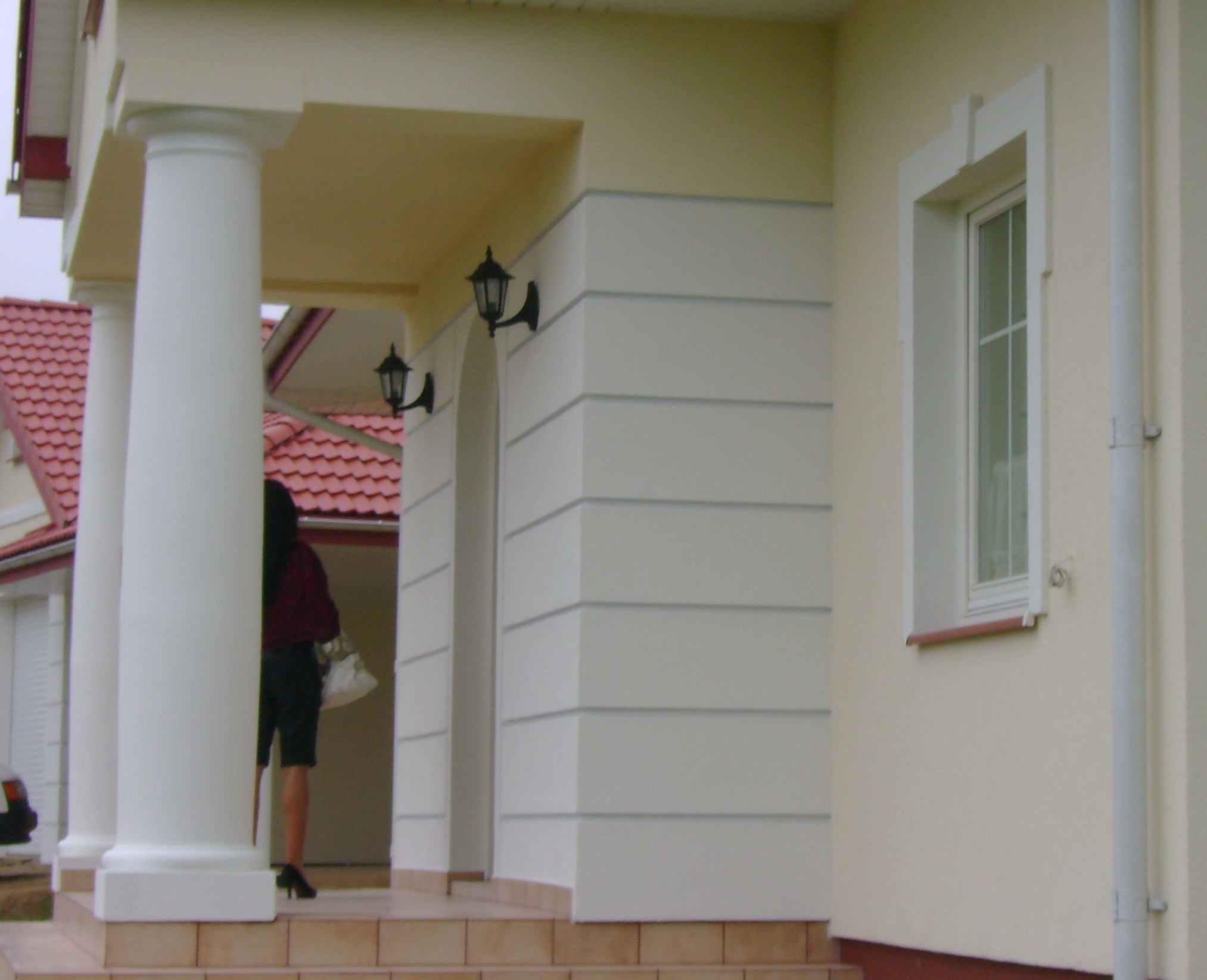 Wejście do domu z kolumnami i dekoracyjną elewacją, widoczne oświetlenie zewnętrzne i okno z ozdobną ramą.